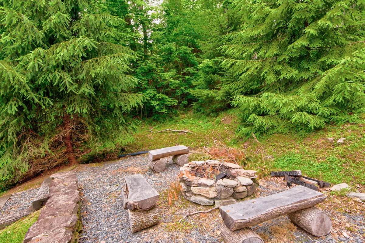 A cozy fire pit area encircled by wooden benches, with surrounding trees creating a secluded, welcoming atmosphere. The scene is ideal for storytelling, making s'mores, and enjoying the warmth of a fire under the night sky.