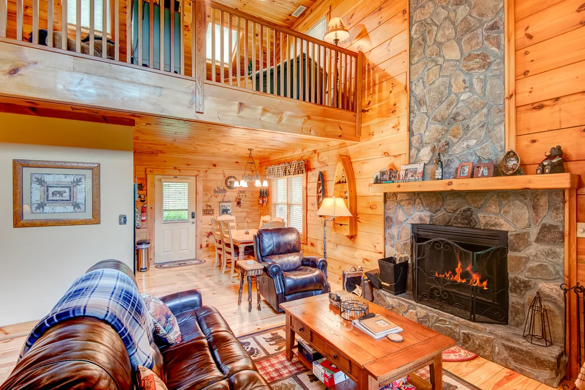 A warm, open-plan interior showcasing a stone fireplace, comfortable leather sofa, visible dining area with wooden table and chairs, rustic wooden beams, and a staircase leading to a loft. The space is bathed in soft natural light, creating an inviting atmosphere for gatherings.