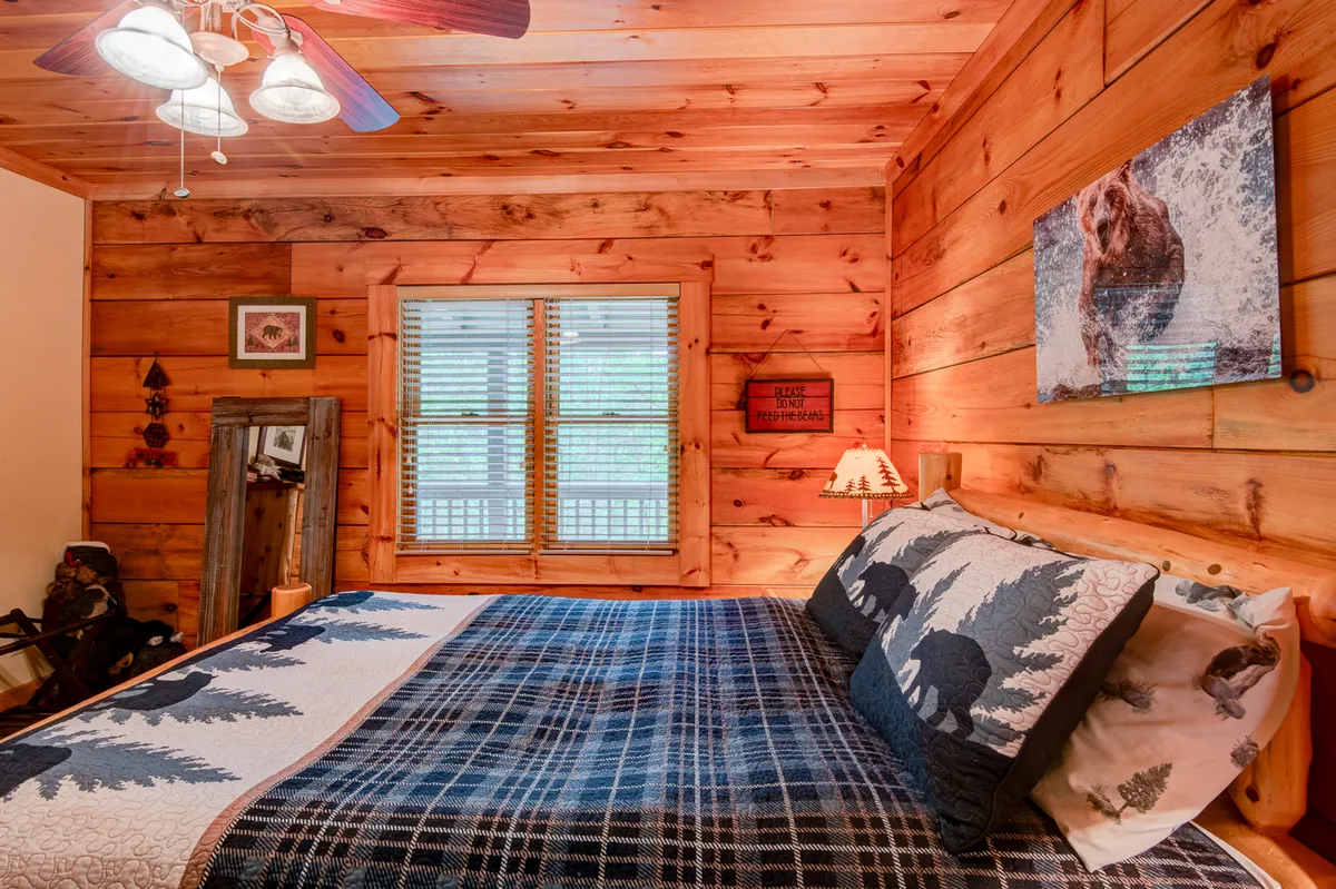 A snug bedroom nook with wooden walls, a bed topped with a patterned quilt, a window with blinds letting in soft light, and bear-themed artwork above the headboard. The mood is peaceful and distinctly cabin-inspired, suggesting a private retreat within the home.