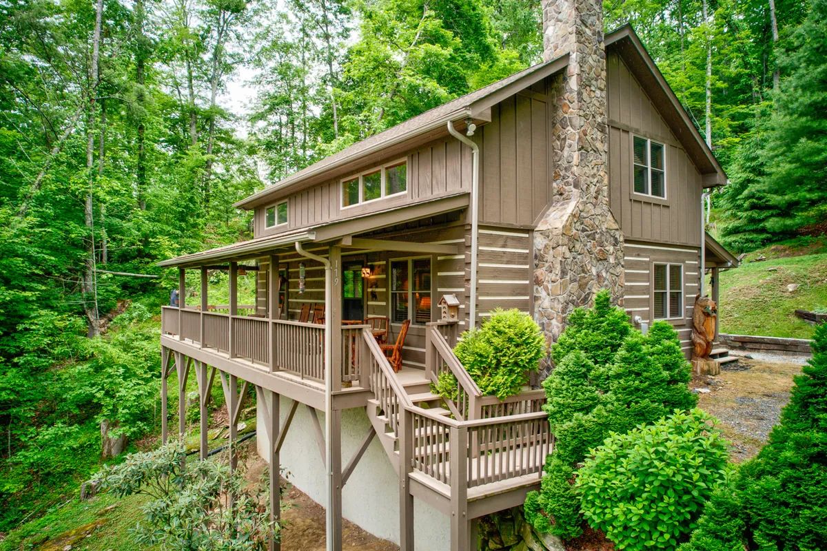 This exterior image showcases the inviting wooden deck of Kaylee's Kabin, the classic log cabin structure, a handsome stone chimney, and abundant lush greenery surrounding the property. The scene suggests a tranquil mountain retreat perfect for relaxing outdoors, with plenty of space for dogs to safely enjoy the fresh air.