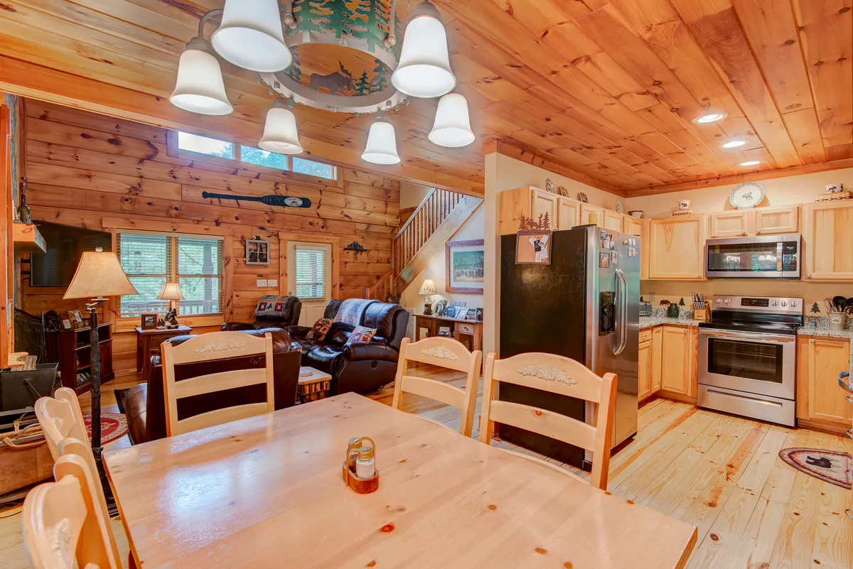 A well-lit, open-concept space that connects the dining table, kitchen appliances, and living area seating. The wood-paneled walls, ample lighting, and visible kitchen amenities reinforce a sense of togetherness and functionality.