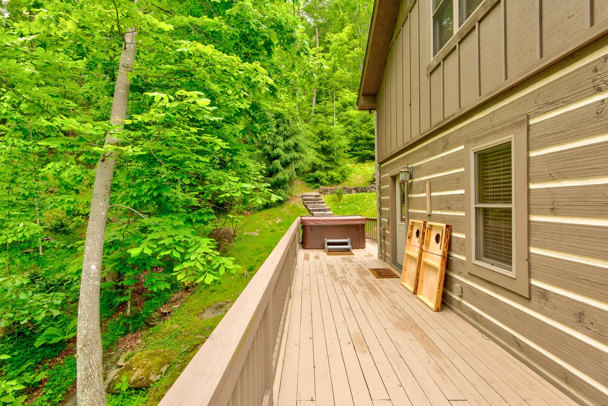 A serene exterior shot of a spacious wooden deck featuring a private hot tub and steps leading into green surroundings. The house's exterior is visible, along with ample room for gathering or relaxing. The hot tub is a focal point, inviting guests to unwind after outdoor adventures.