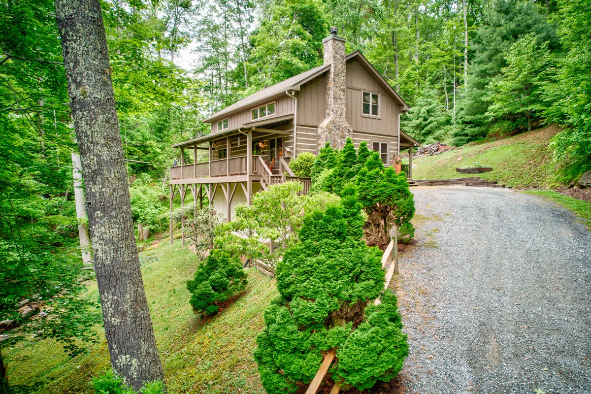 This image features the house surrounded by lush greenery, a gravel driveway leading up to the entrance, and an inviting porch. The setting hints at the peacefulness and accessibility of outdoor experiences right outside the cabin, with ample space for families or friends to gather and enjoy nature.