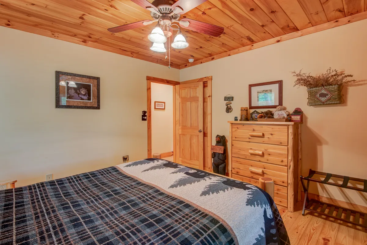 A warm, inviting bedroom featuring a wooden ceiling, plaid patterned bedding on a large bed, a wooden dresser, and framed artwork on the walls. Sunlight gently filters through the window, enhancing the cozy atmosphere and emphasizing the restful nature of the space.