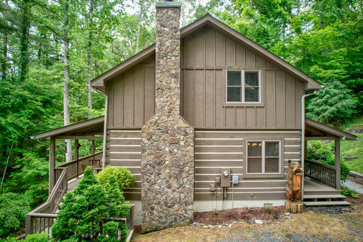 A welcoming view of the wooden deck featuring a stone chimney, landscaped garden, and a carved wooden bear, all set against a backdrop of trees. The outdoor gathering space highlights opportunities for evening bonfires, morning coffee, and soaking in nature—activities central to mountain getaways.