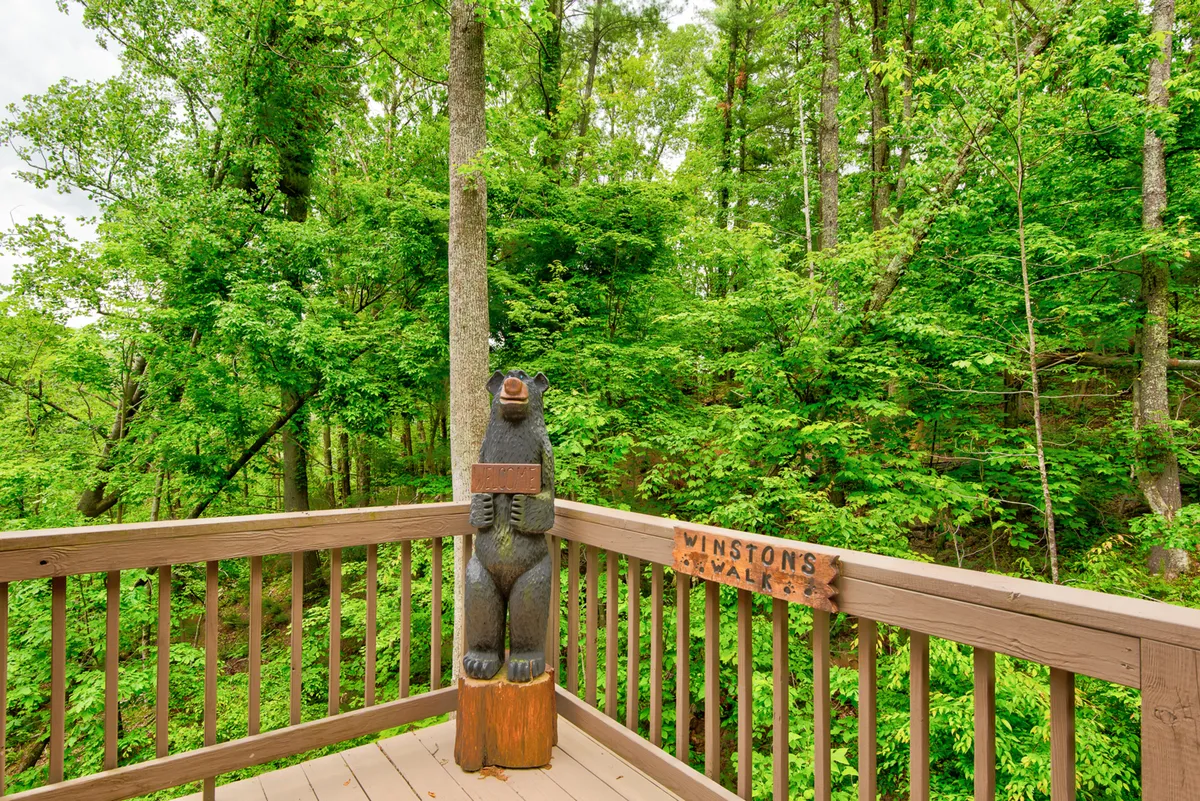 A playful view of the wooden deck with a wooden bear statue and deck railing, overlooking lush green trees. The railing is prominent, hinting at the secure, enclosed nature of the deck—ideal for dog owners concerned about safety.