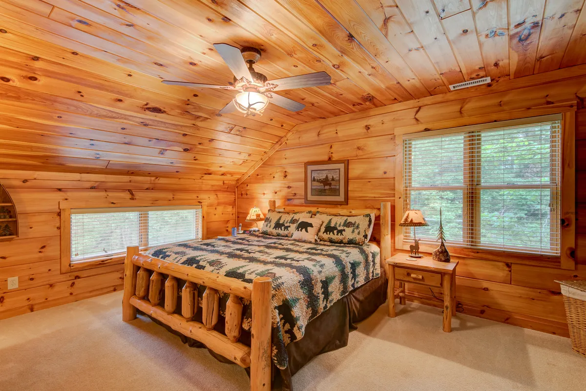 A cozy bedroom with a rustic log bed adorned with bear-themed bedding, flanked by wooden nightstands and panorama windows that look out to nature. The setting feels both tranquil and inviting, ideal for quiet relaxation or waking up to a scenic view.