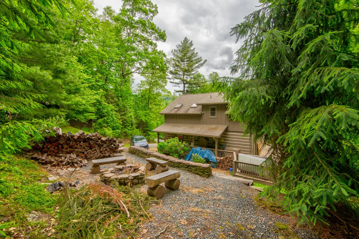 This image shows the house, a woodpile, and a stone seating area in the yard, with a car parked nearby. The setting is welcoming, featuring both the home and designated space for outdoor gatherings, suggesting activities like fire pit evenings and playtime for kids.
