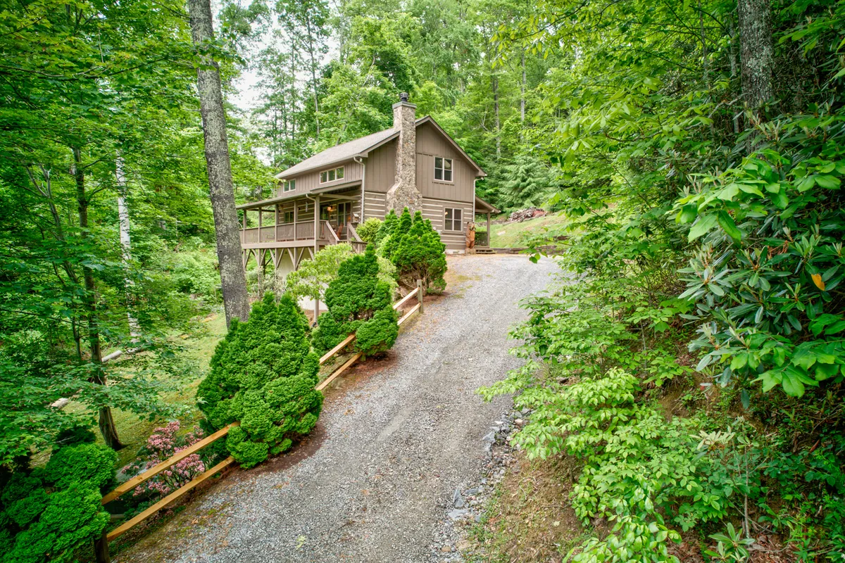 The image showcases the charming log cabin set amid tranquil trees, with a welcoming driveway leading up to the home. Gated decks wrap around the exterior, inviting guests to relax and take in the peaceful, natural surroundings. Lush landscaped shrubs and the quiet forest backdrop create a serene, private atmosphere ideal for relaxation.