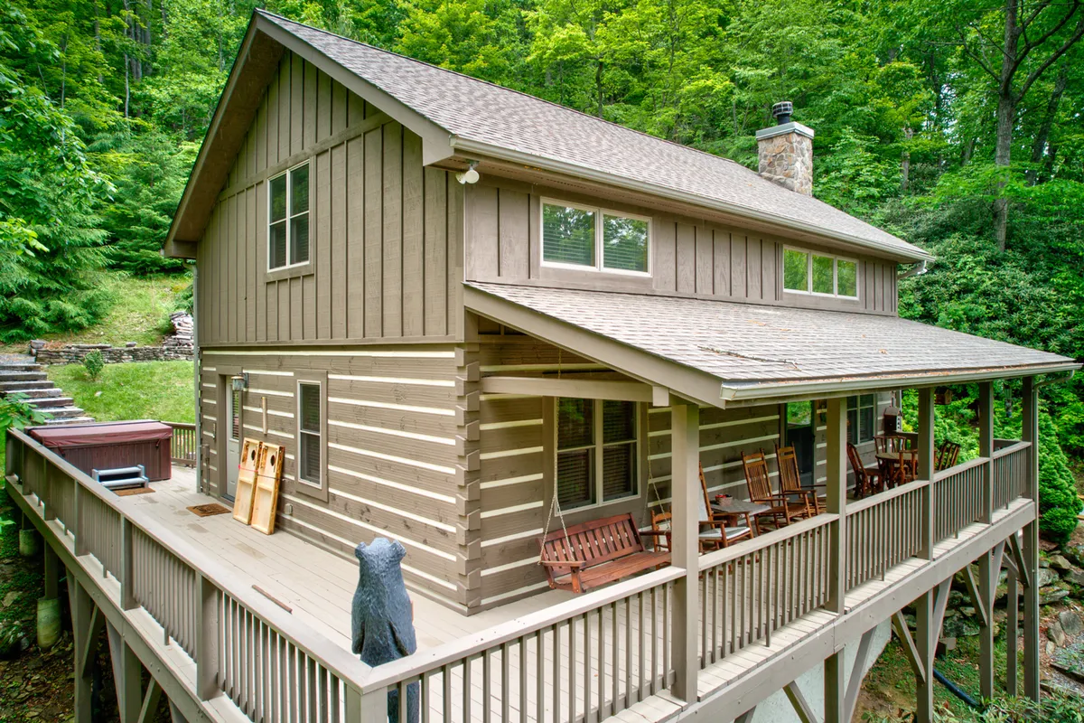 This image features the exterior of Kaylee's Kabin, with wooden rocking chairs lined up on the porch, a prominent stone chimney, and lush natural greenery enveloping the cabin. The tranquil setting invites guests to imagine relaxing after a day of mountain adventure, with the porch providing the perfect spot to soak in the High Country atmosphere.